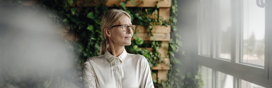 A mature professional woman stands in front of a vine-covered trellis and looks thoughtfully out a glass window. 